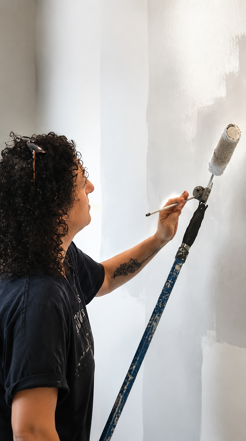 A person with curly dark hair applies white paint to a wall using a long-handled paint roller and brush. The person wears a dark navy T-shirt, and the wall being painted is a light off-white.
